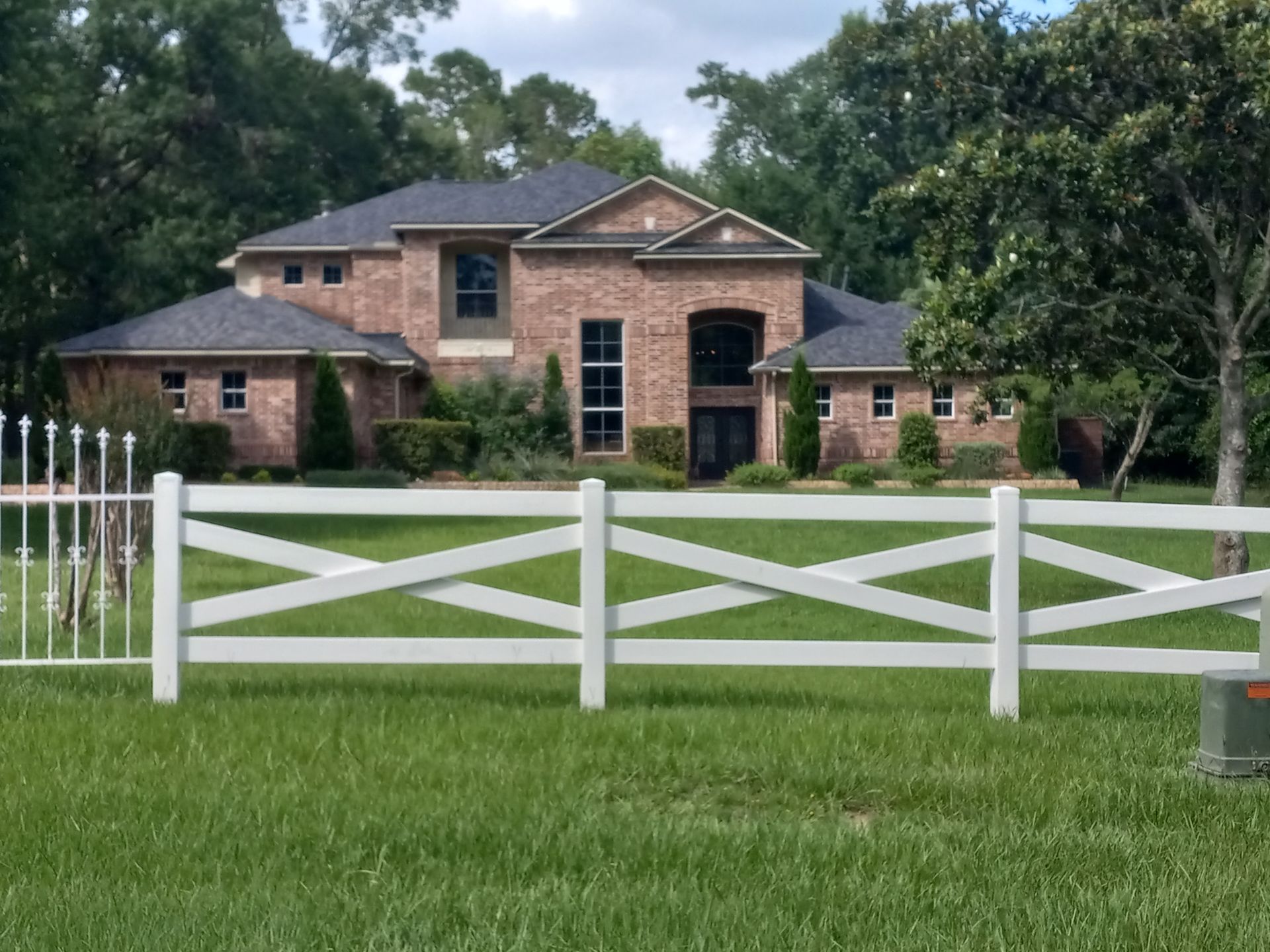 Brick two-story house with dark roof, white crisscross fence in front of green lawn and trees.