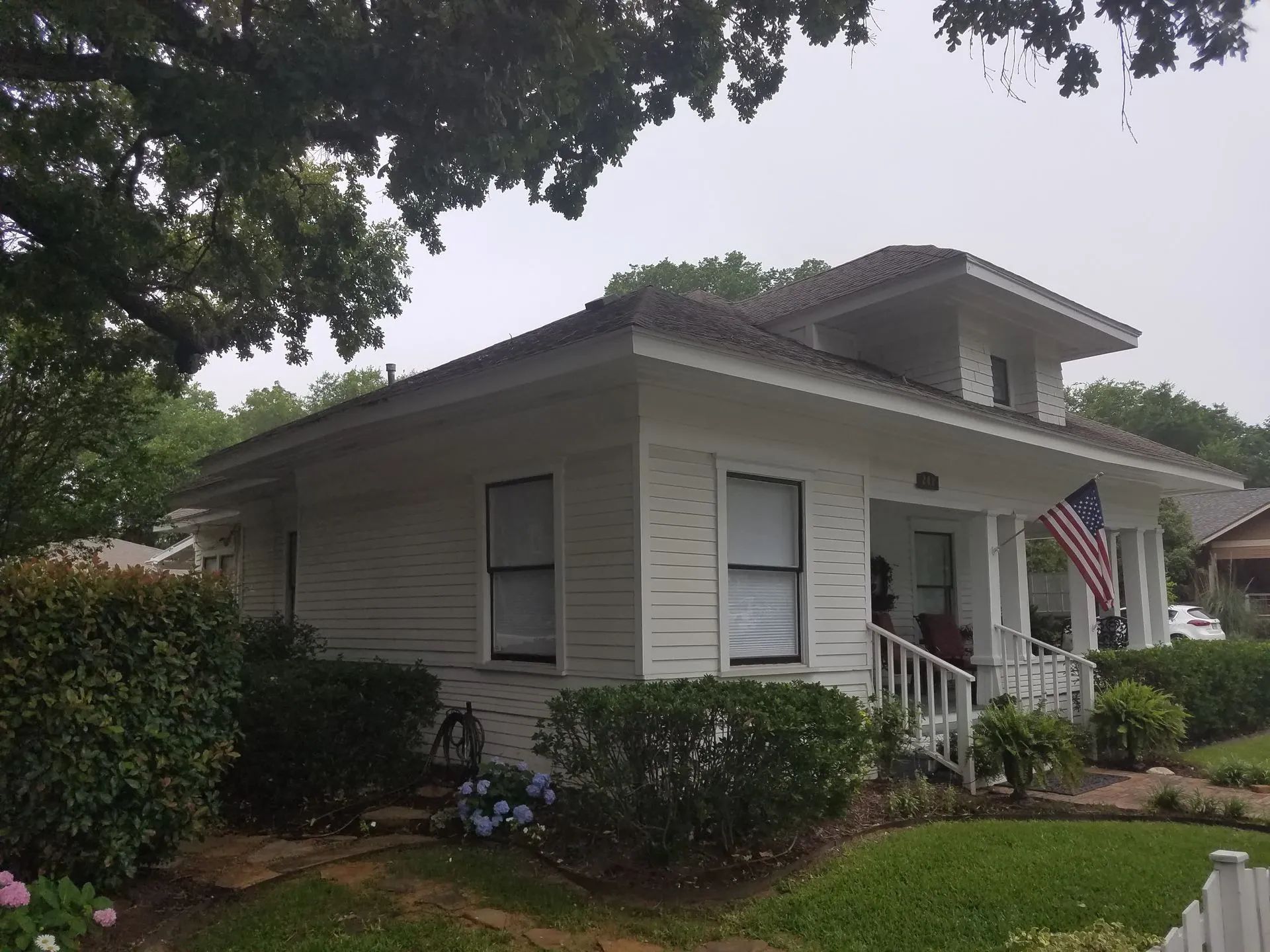 White Craftsman house with a porch, American flag, and lush greenery in front.