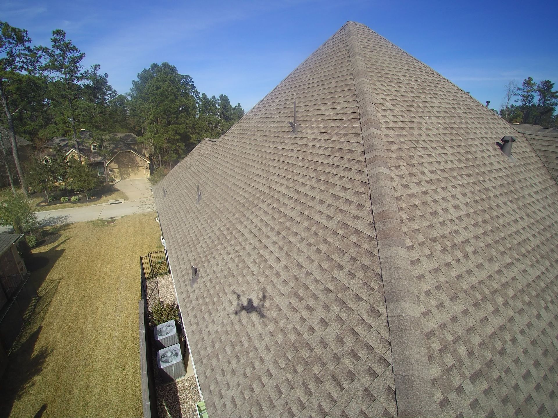 Brown shingle roof on a house, viewed from above, with blue sky and trees in the background.