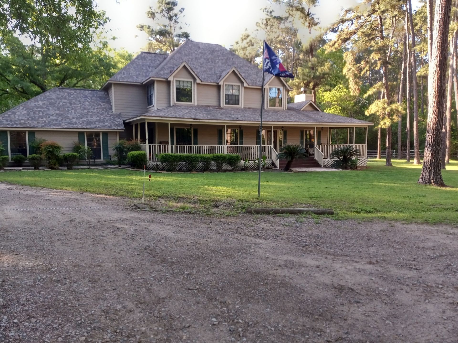 Tan two-story house with porch and large lawn, trees in the background, gravel driveway.