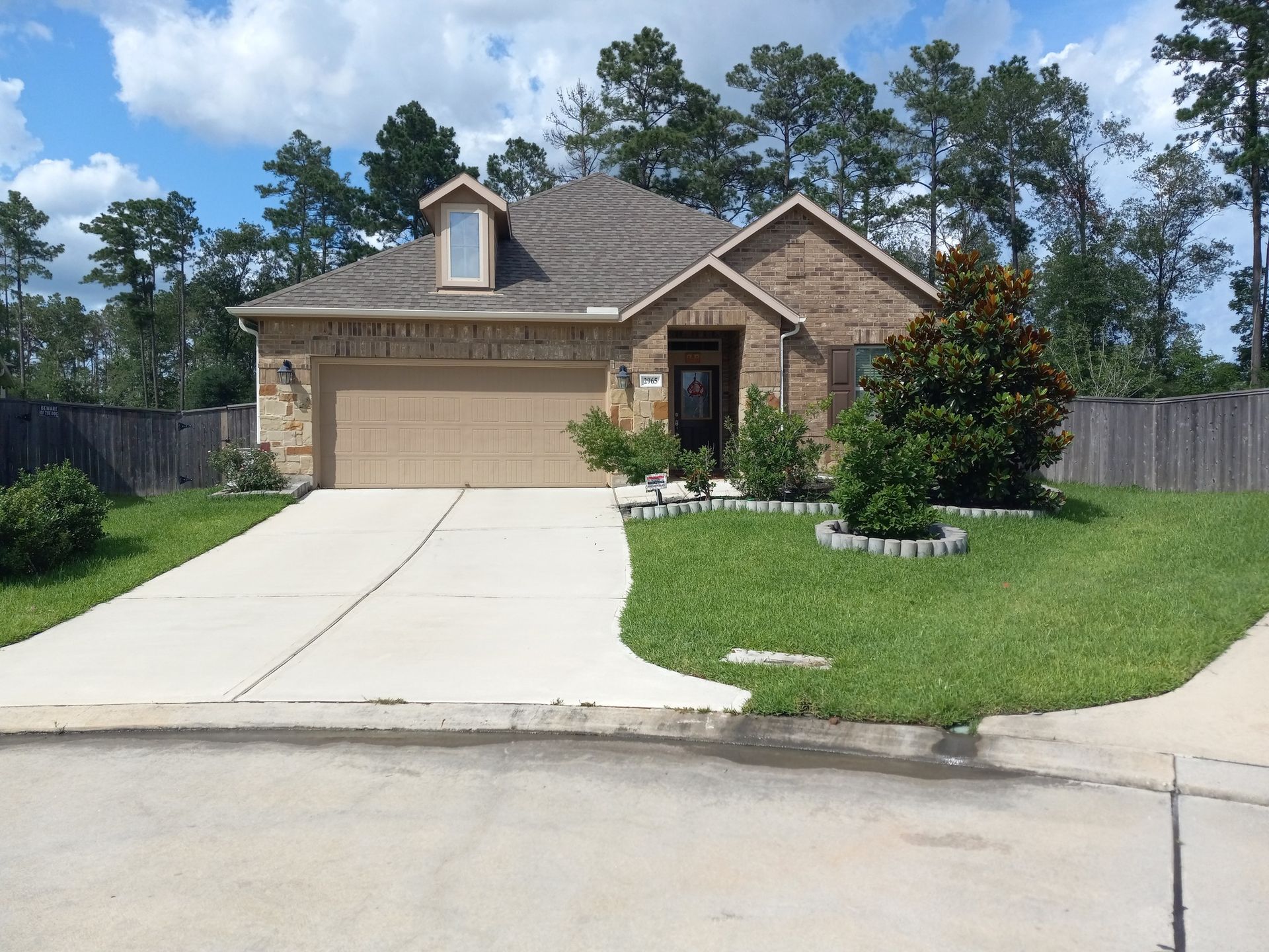 Tan brick house with a dormer, attached garage, and well-manicured lawn. Blue sky with trees in background.
