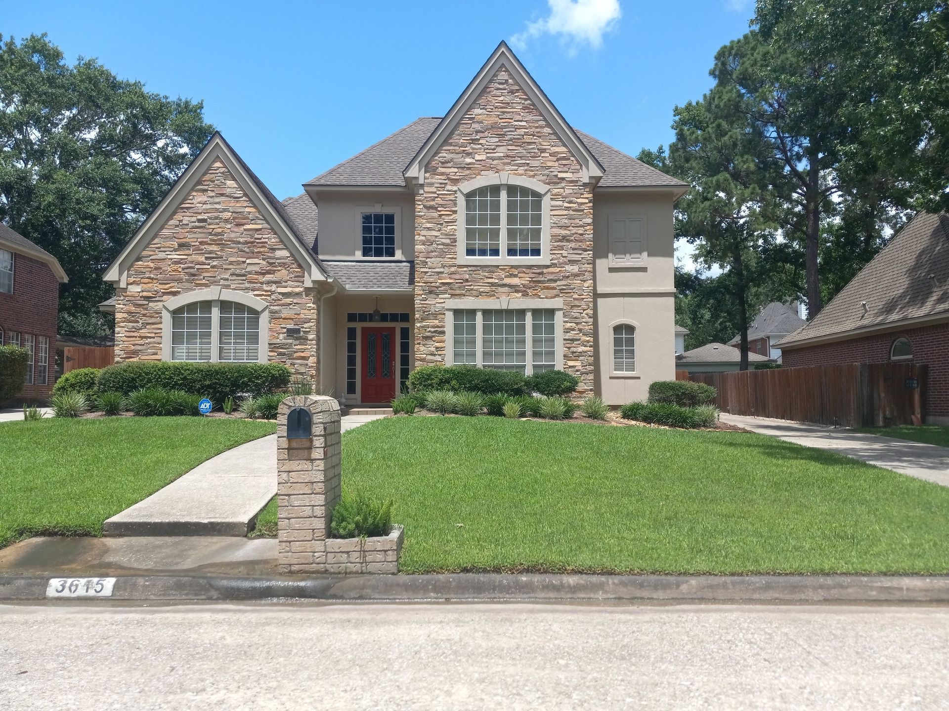 House with stone and stucco facade, red door, manicured lawn, trees.