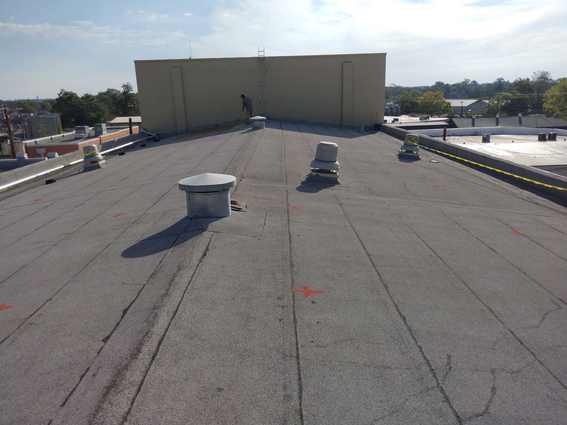 A flat, gray commercial rooftop with vents and a person working near a beige building in daylight.