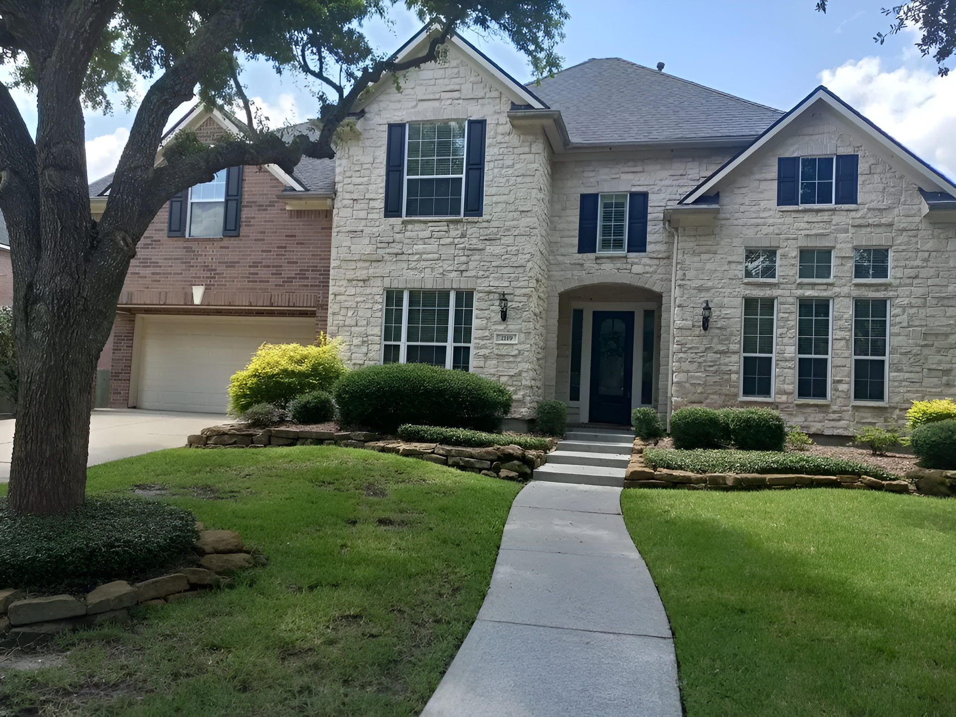 Stone-clad two-story house with a dark door, black shutters, and a curved concrete walkway. Lush green lawn.