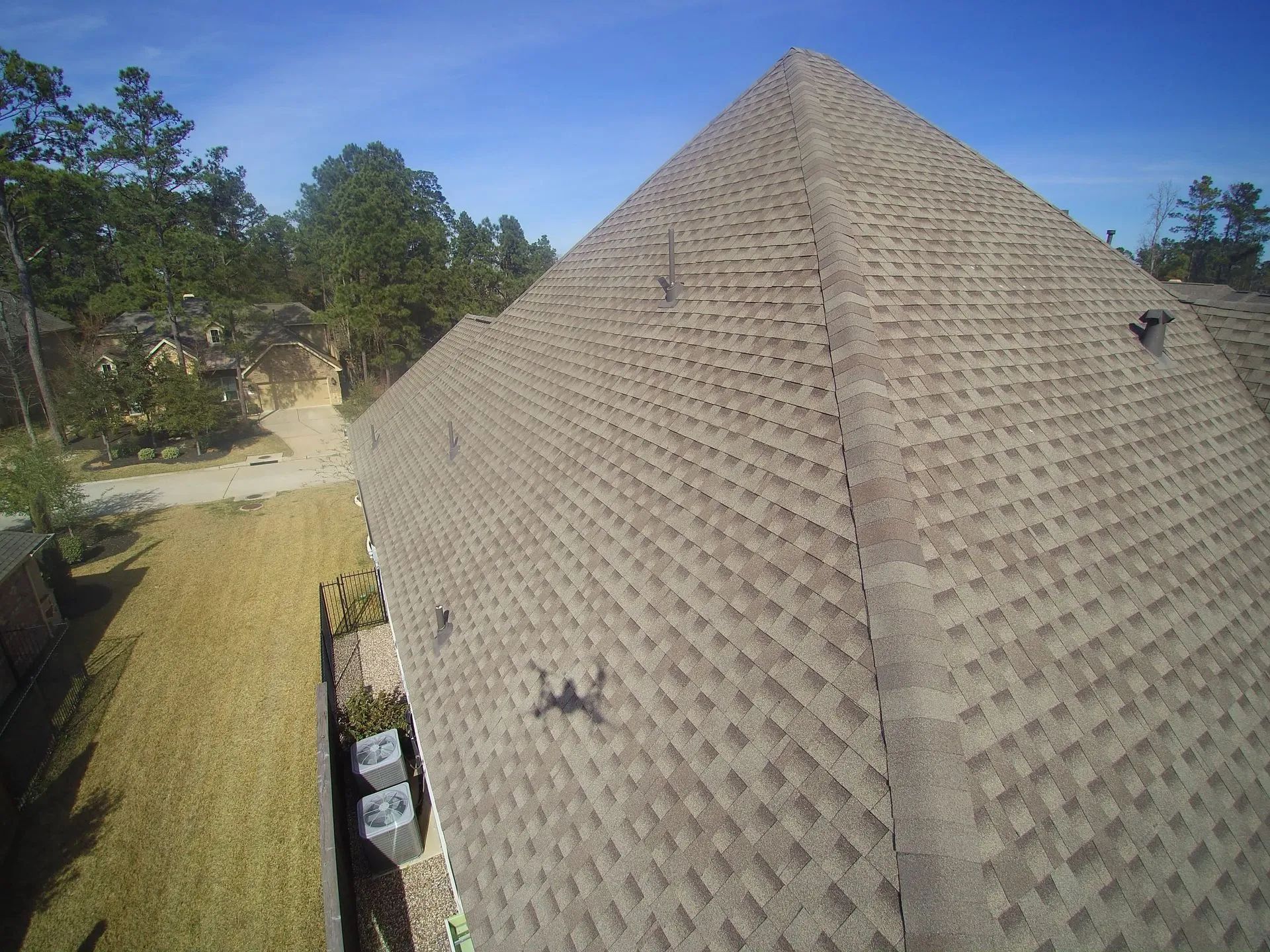High-angle view of a brown shingled roof on a house, with a grassy yard, trees, and blue sky.