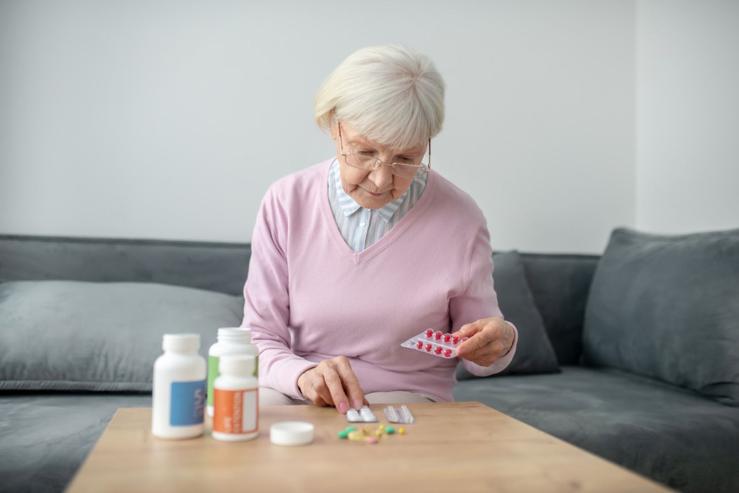 Elderly woman with glasses, sorting pills on a table, seated on a couch.