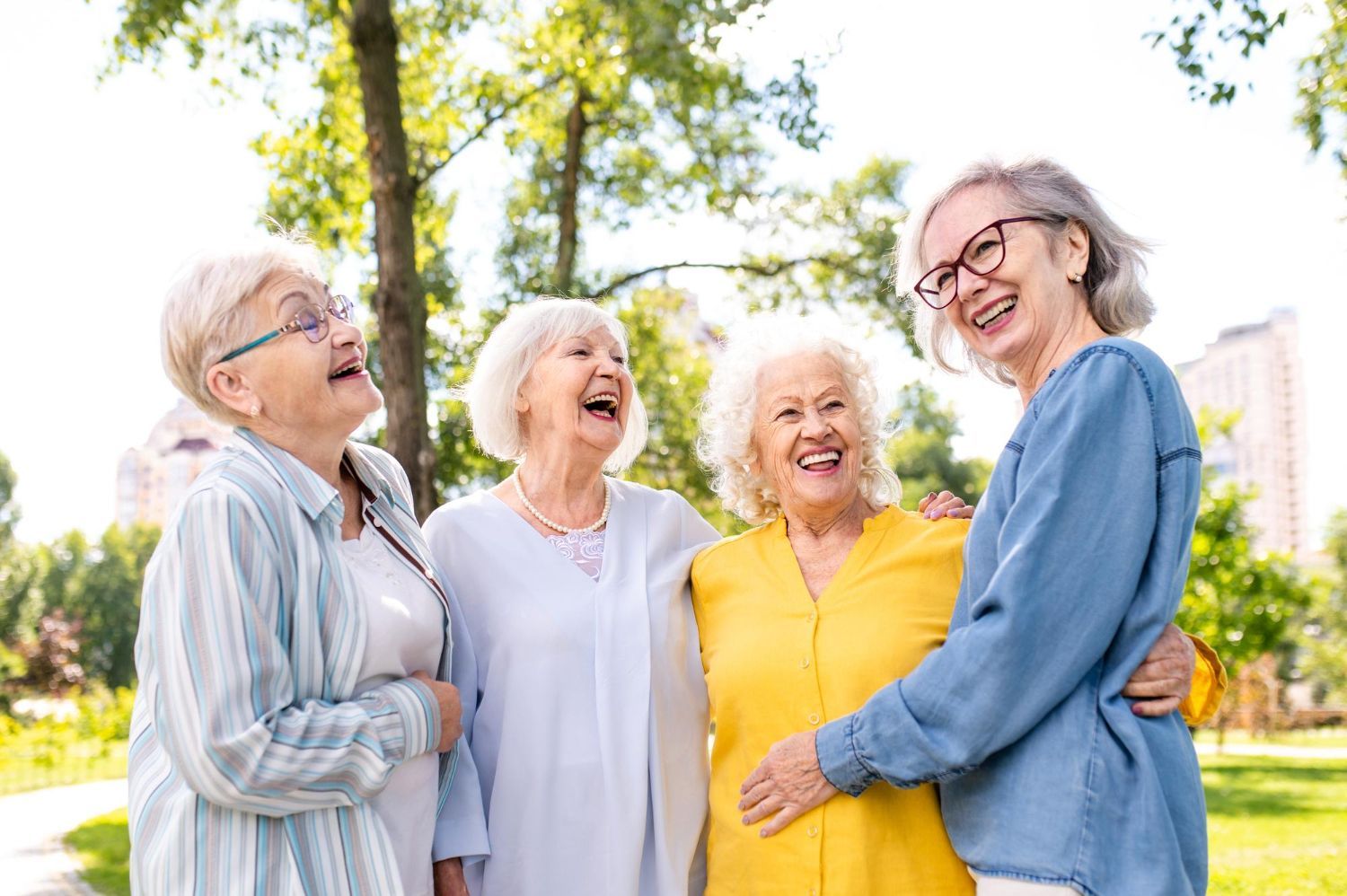 Four elderly women laughing together in a park, embracing and enjoying the outdoors.