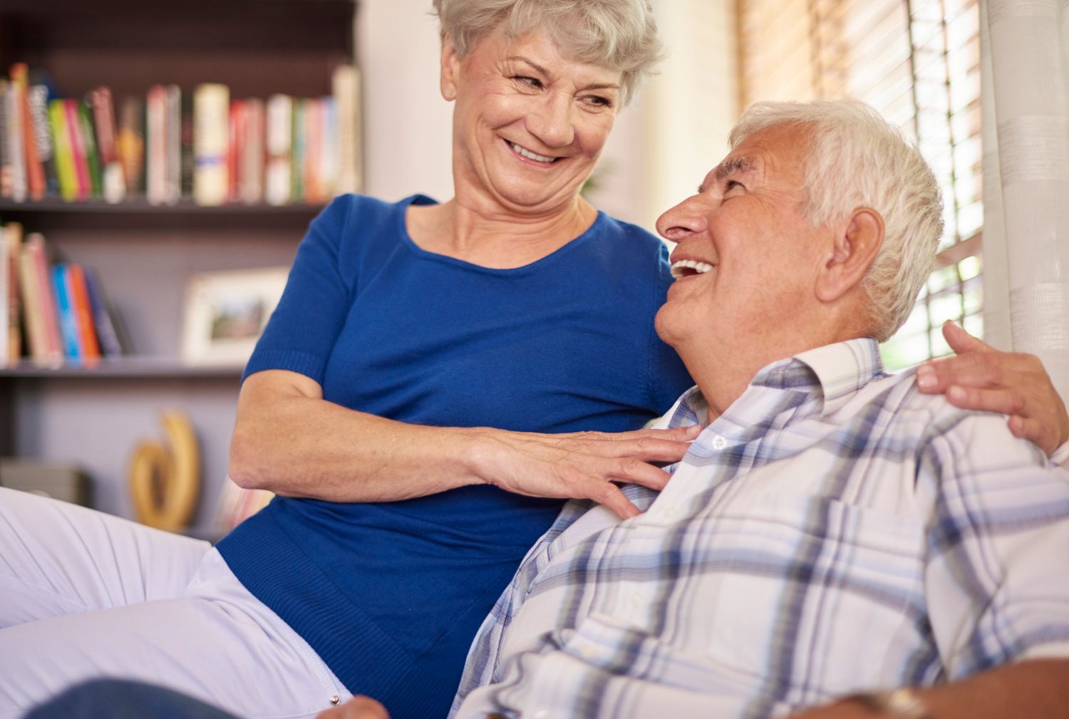 Elderly couple smiling at each other, woman's arm around man sitting on couch in living room.