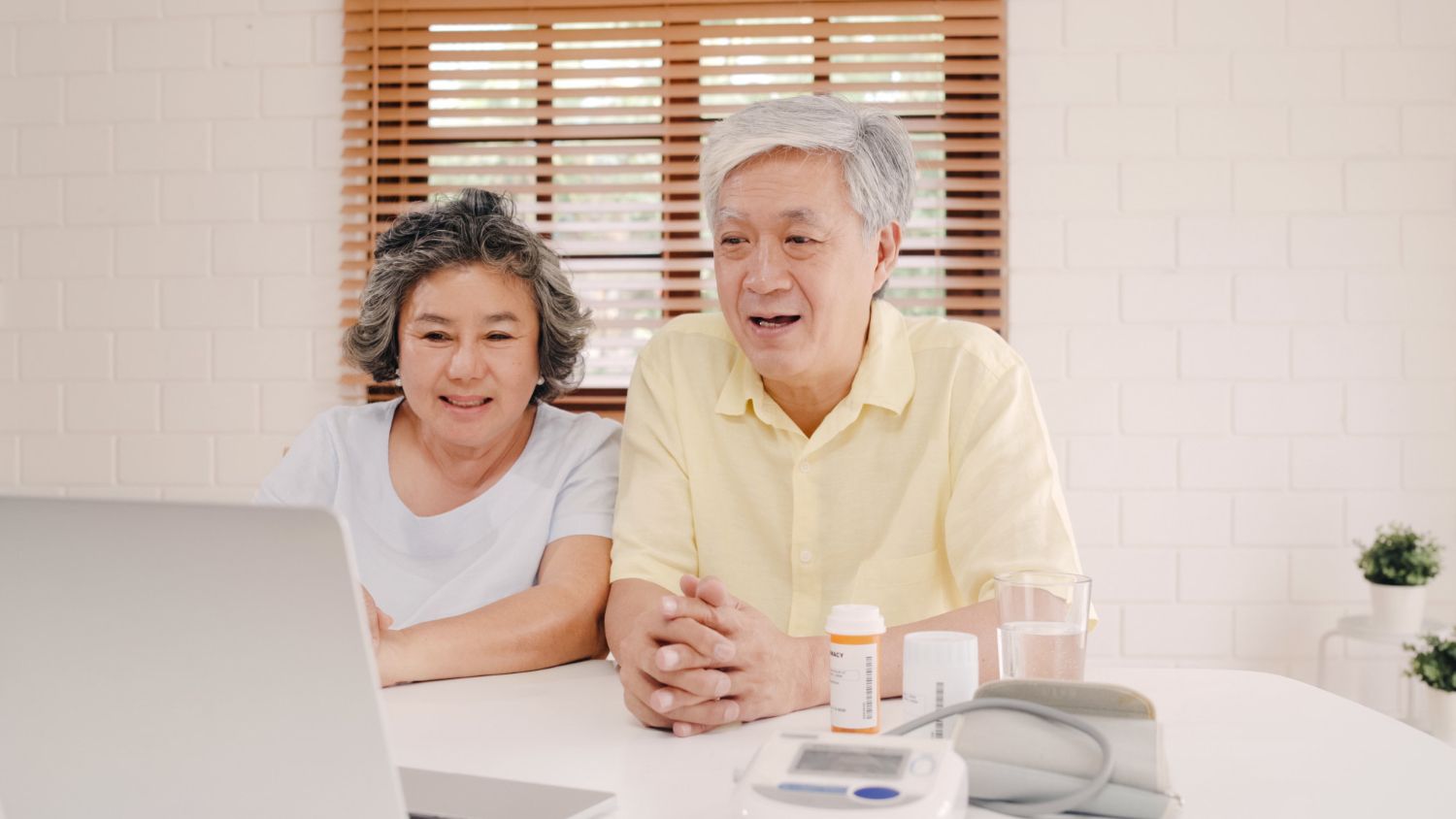 Elderly couple using a laptop, possibly for a telehealth appointment. White table, pills, blood pressure monitor. Happy expressions.