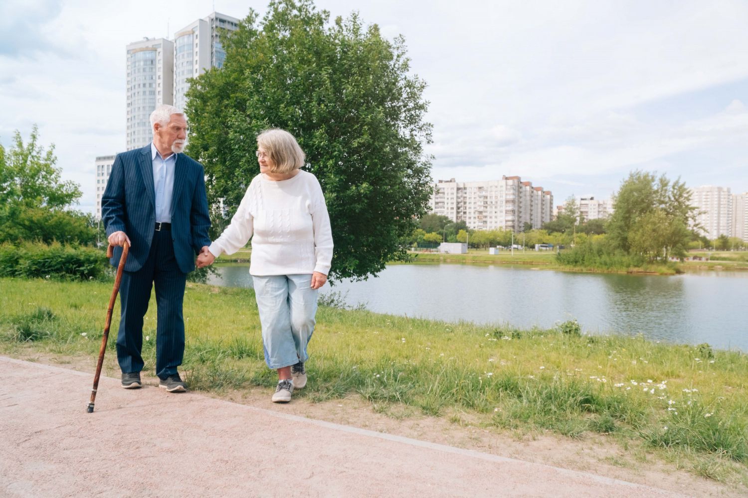 Elderly couple walking, holding hands, along a path by a lake. Man uses a cane.