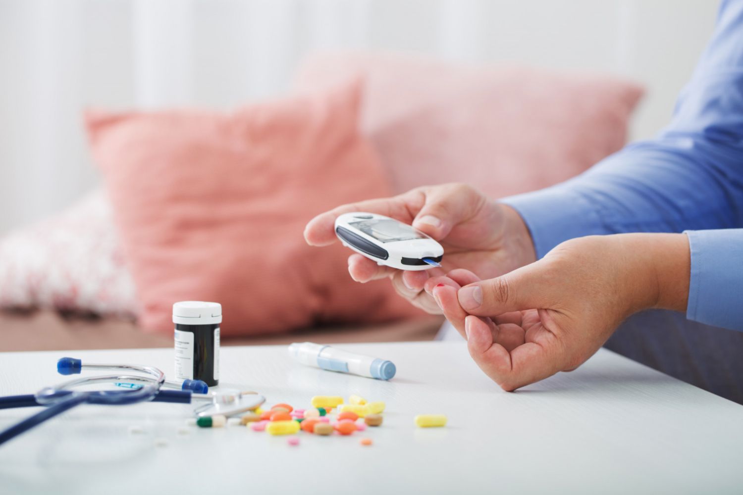 Person testing blood sugar with a glucometer, pills, and medical supplies on a white table.