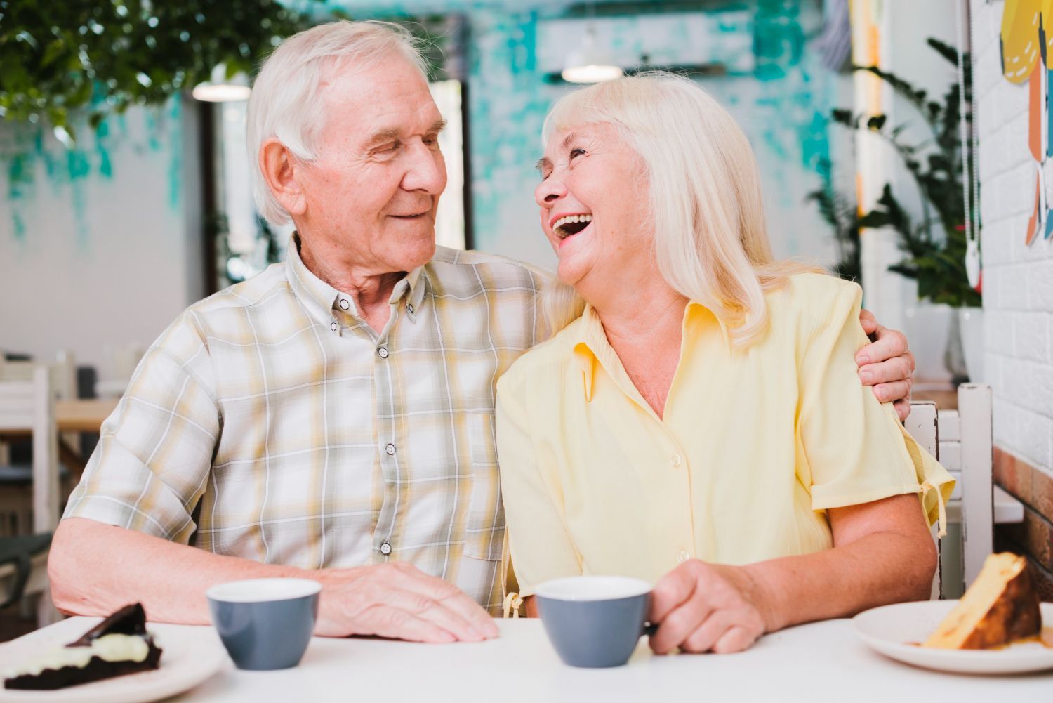 Senior couple laughing together at a table in a cafe, enjoying coffee and cake.