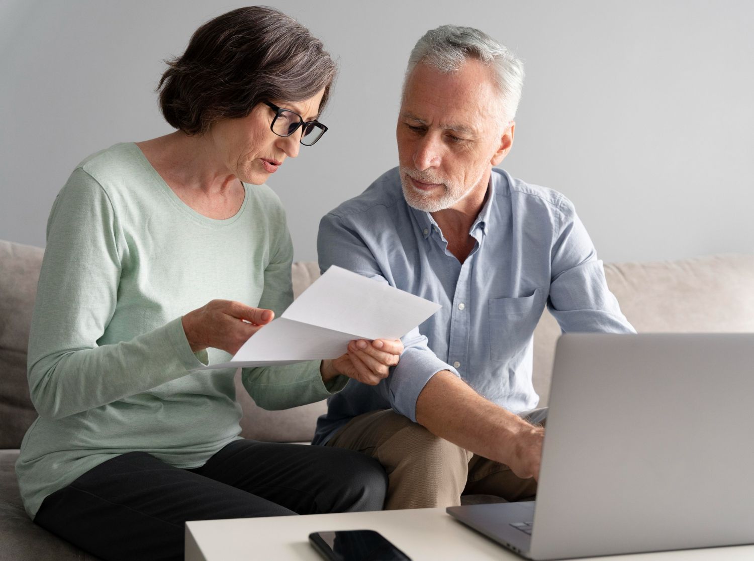 An older couple reviews papers while using a laptop on a couch. The woman wears glasses, the man is looking at the screen.