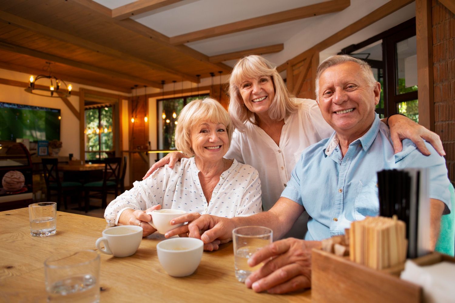 Three smiling seniors at a bar; two women and a man. One woman has an arm around the man. They are holding cups and glasses.