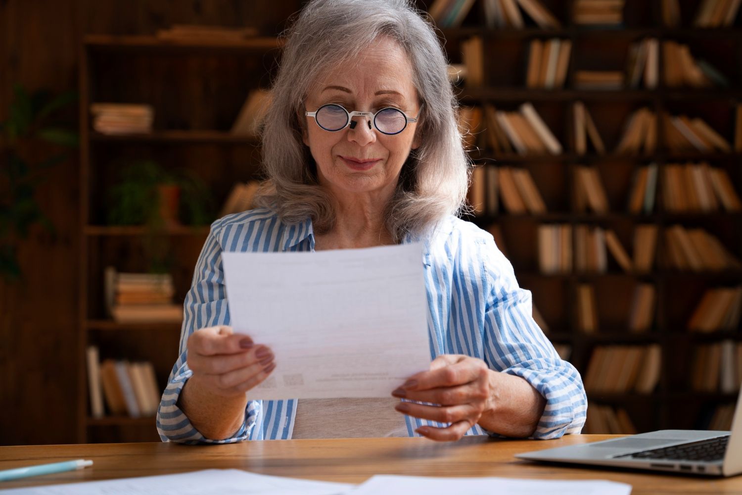 Elderly woman with glasses reads a document at a desk, library books in the background.