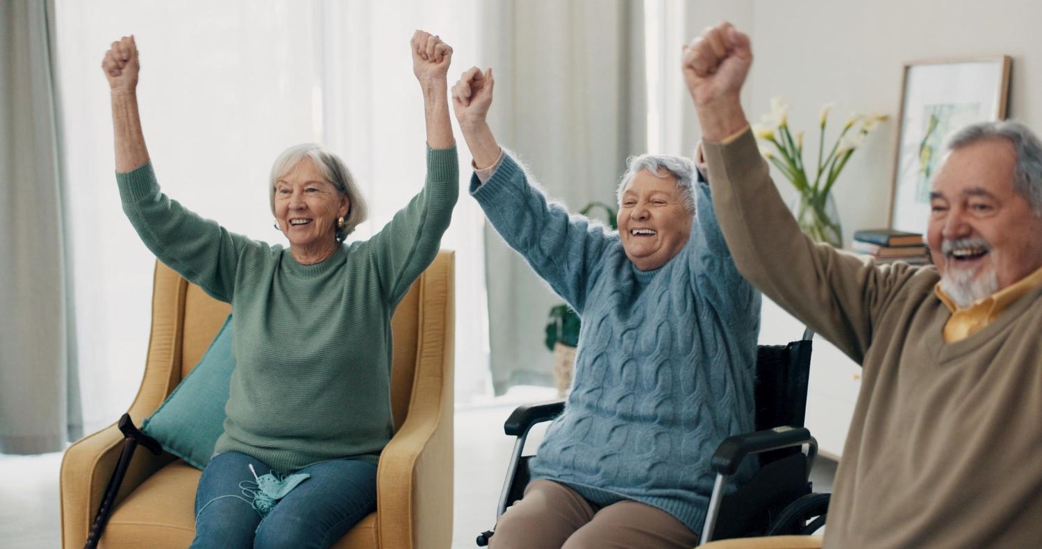 Three smiling seniors raising arms, celebrating in a bright living room; one in wheelchair.
