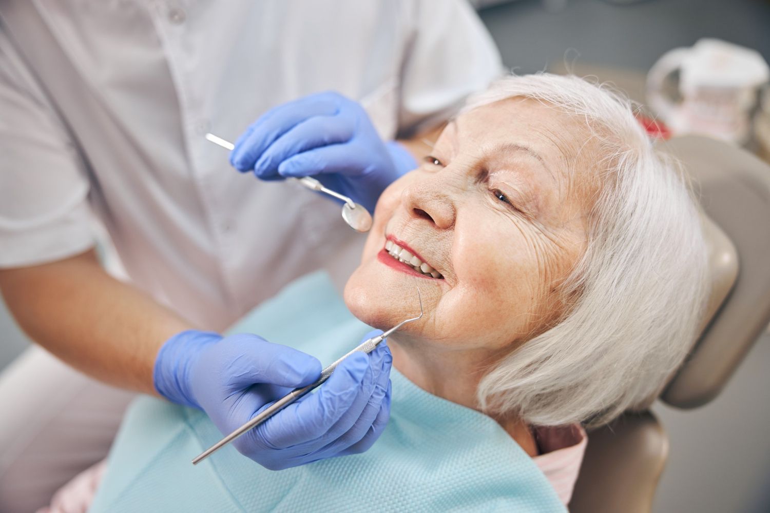 A senior woman smiles while receiving a dental checkup. A dentist in gloves holds tools.