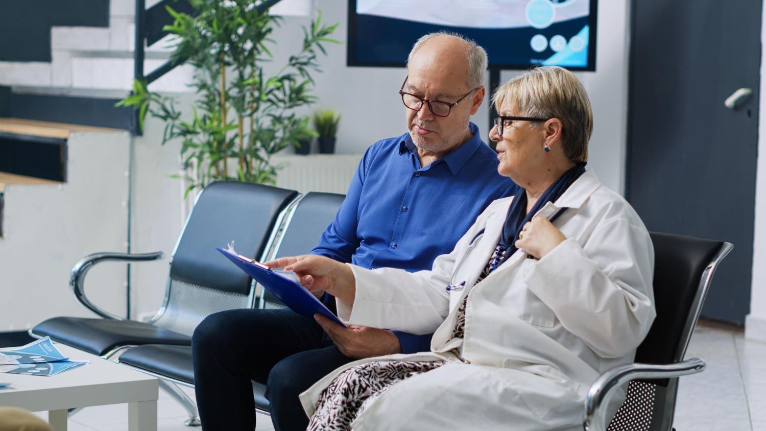 A doctor in a white coat shows a medical chart to an older patient in a waiting room.