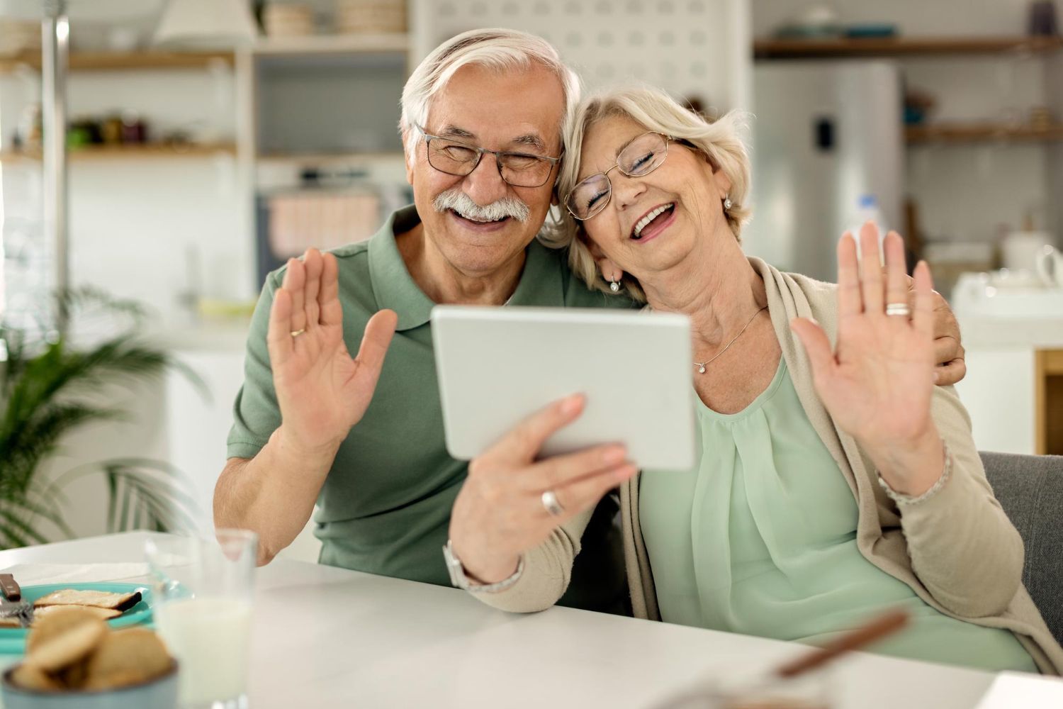 Elderly couple waves during a video call, smiling. They sit at a table in a bright room.