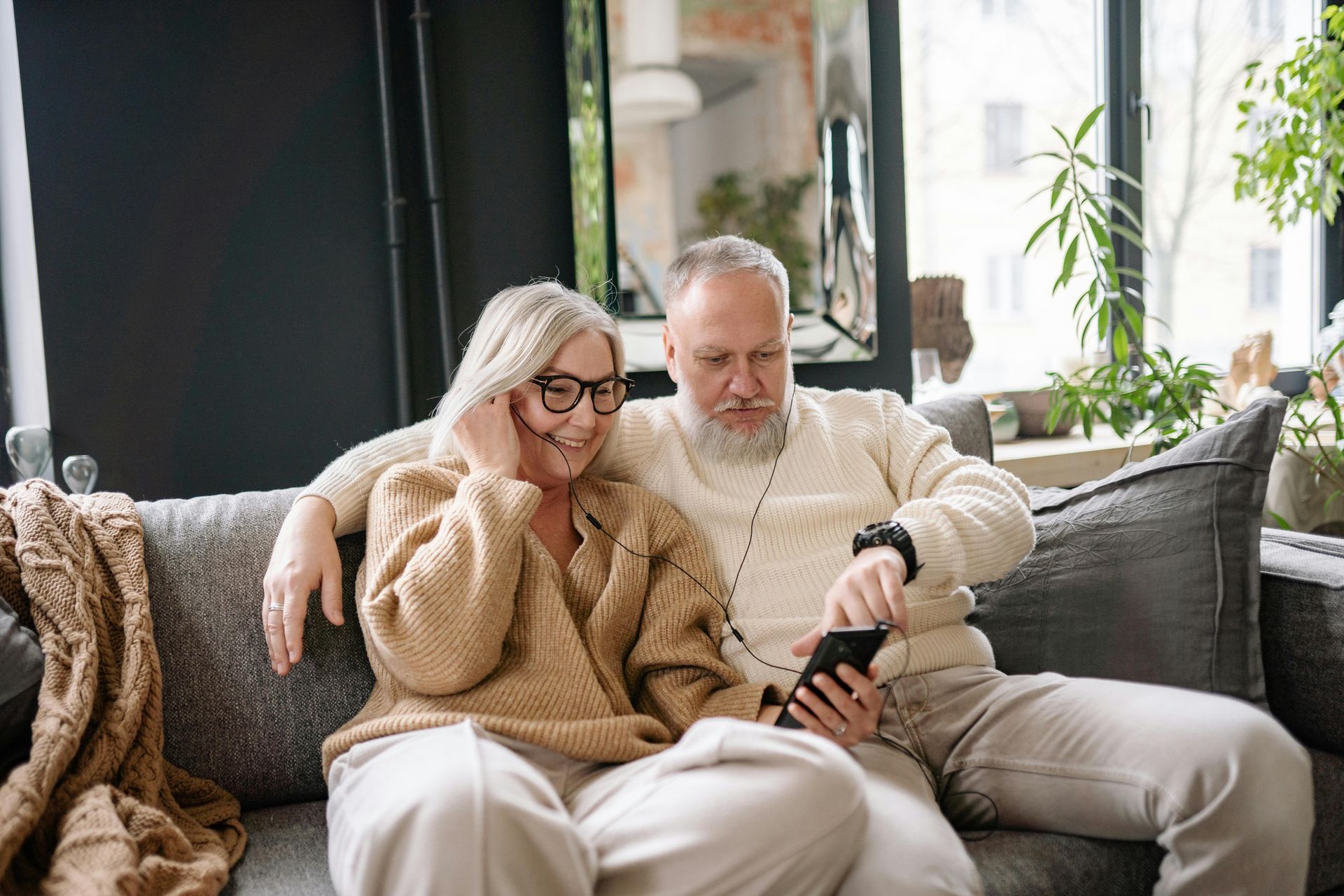 Senior couple sitting on a couch looking at a smartphone, smiling. Cozy living room setting.