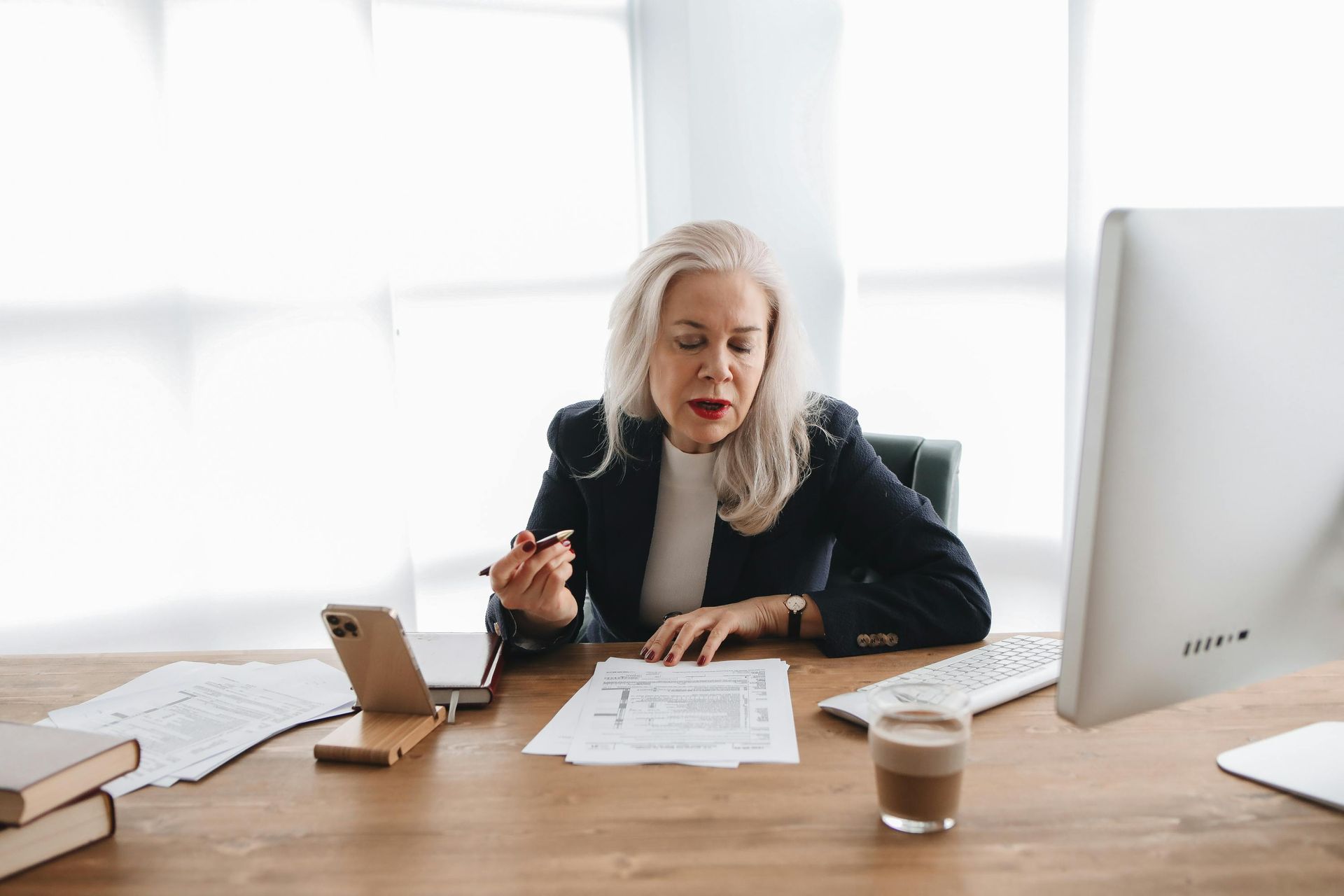 Woman with gray hair in blazer reviews paperwork at desk with computer and phone.