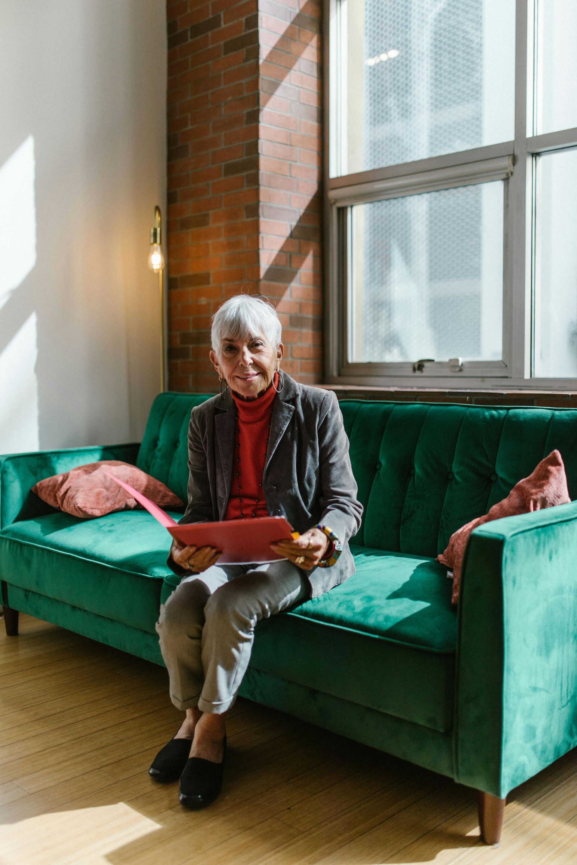 Woman with grey hair smiles, holding a pink folder, seated on a green couch in front of a window.