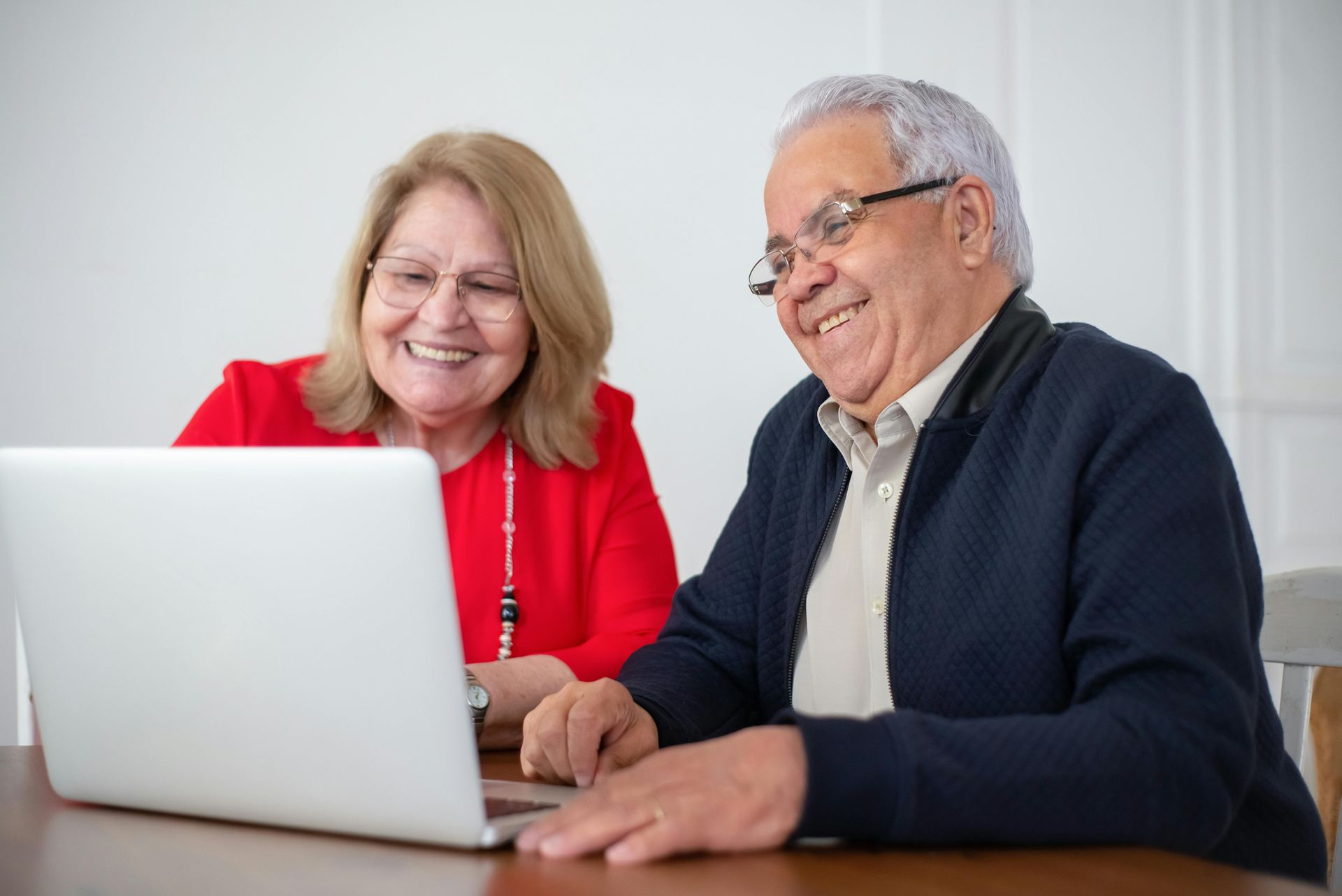 Senior couple smiles while looking at a laptop computer. Woman in red, man in navy. Indoors.