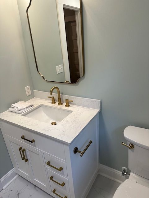 A modern bathroom vanity with a white cabinet, gold hardware, a white sink, a marble countertop, and a gold-rimmed mirror.