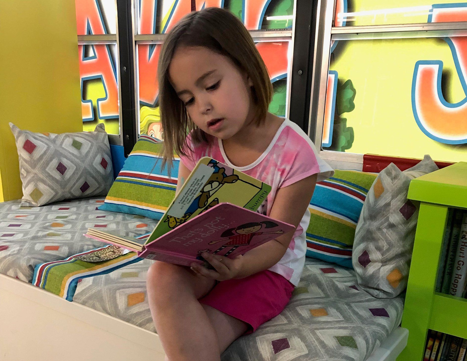 A person sits on a colorful cushioned bench, reading an open book in a bright, play-oriented room.