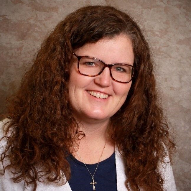 A smiling person with long curly brown hair and glasses, wearing a dark shirt, a light cardigan, and a cross necklace.