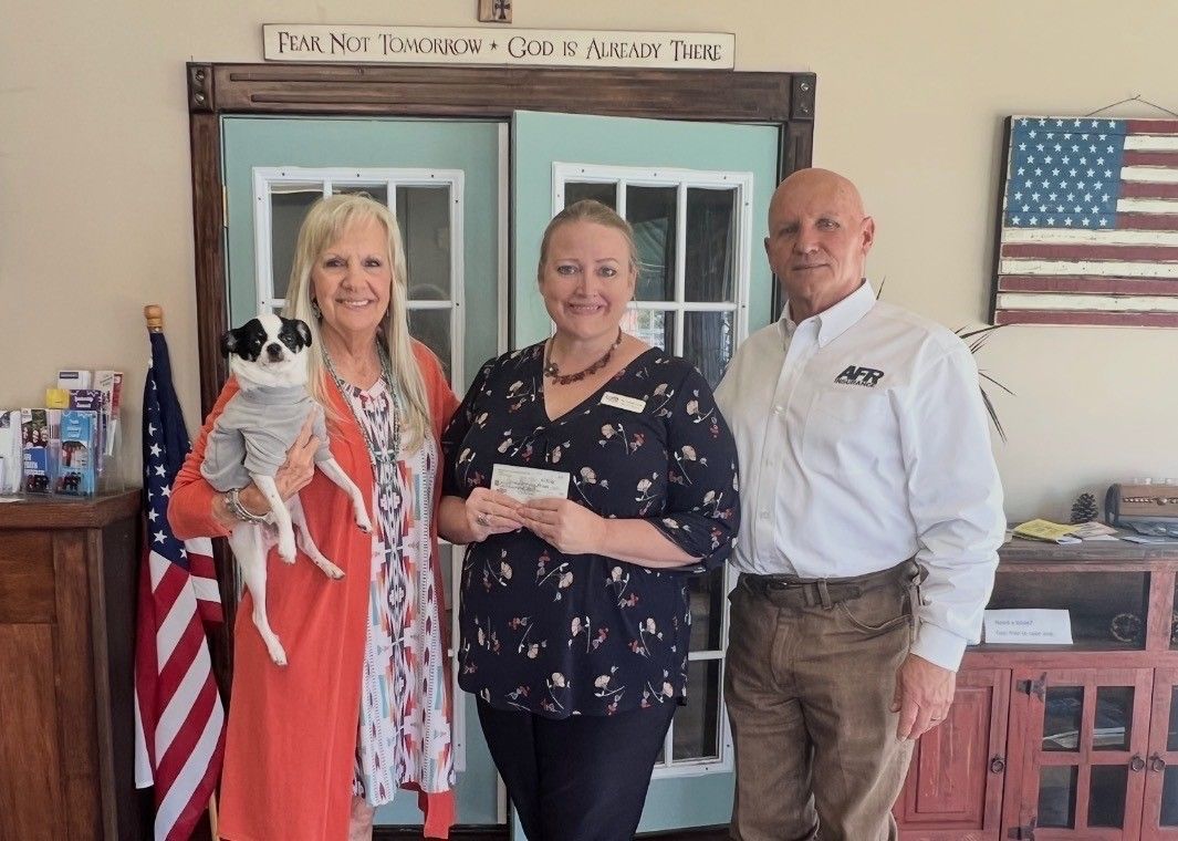 Three people stand indoors with a small dog; one holds an envelope, with an American flag displayed in the background.