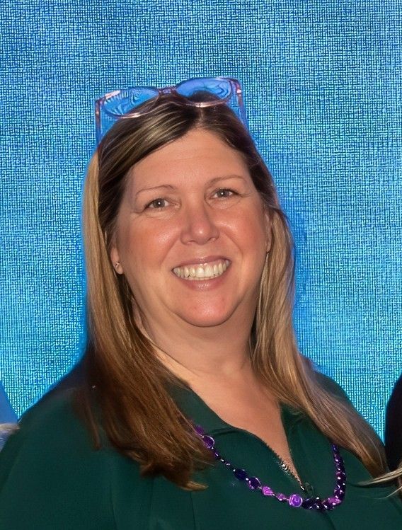 A person smiling, wearing glasses on their head, a dark green top, and a beaded necklace against a blue textured backdrop.