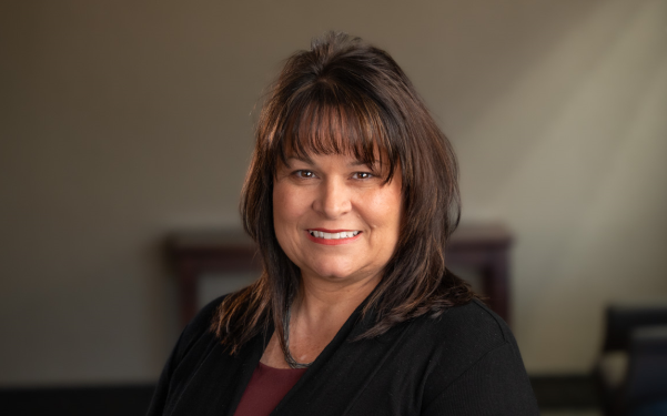 A professional headshot of a smiling woman with shoulder-length brown hair, wearing a black top against a neutral background.