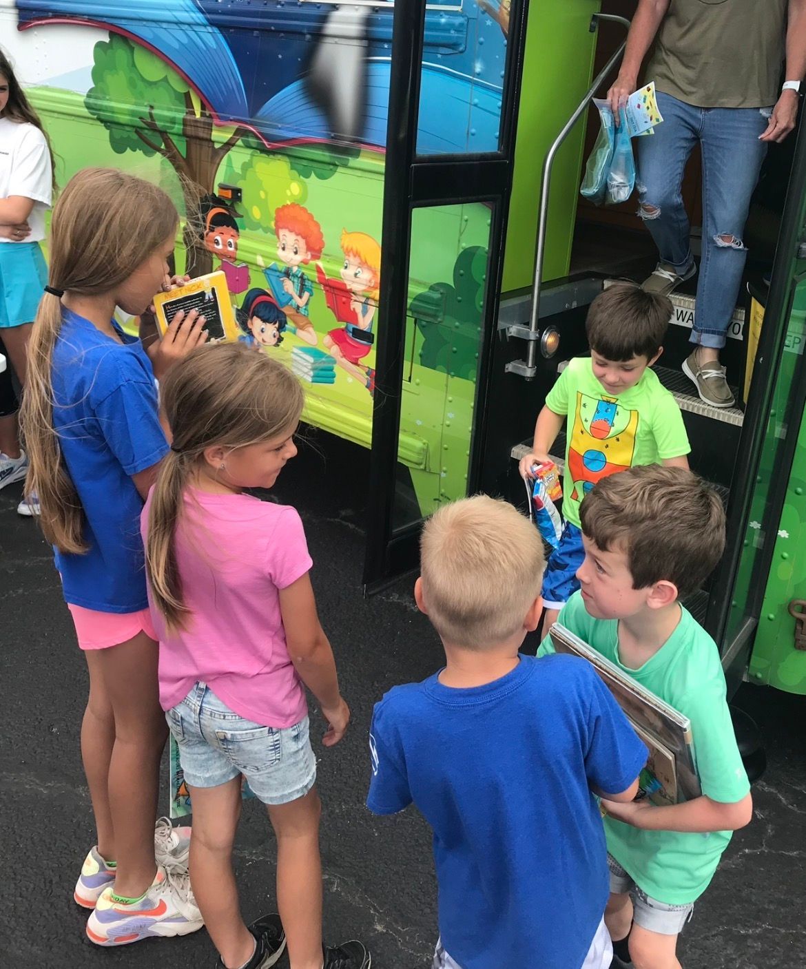 Children gather near the steps of a colorful, mural-painted bus while a person prepares to hand out items from inside.