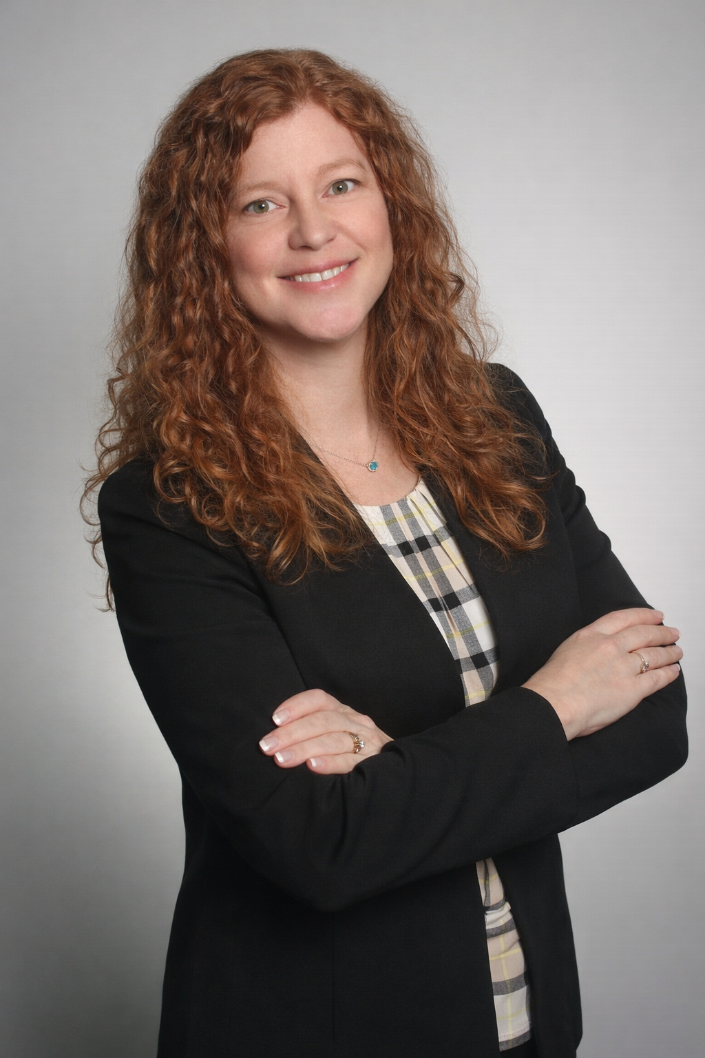 A person with wavy reddish-brown hair smiling with arms crossed, wearing a black blazer over a plaid top.