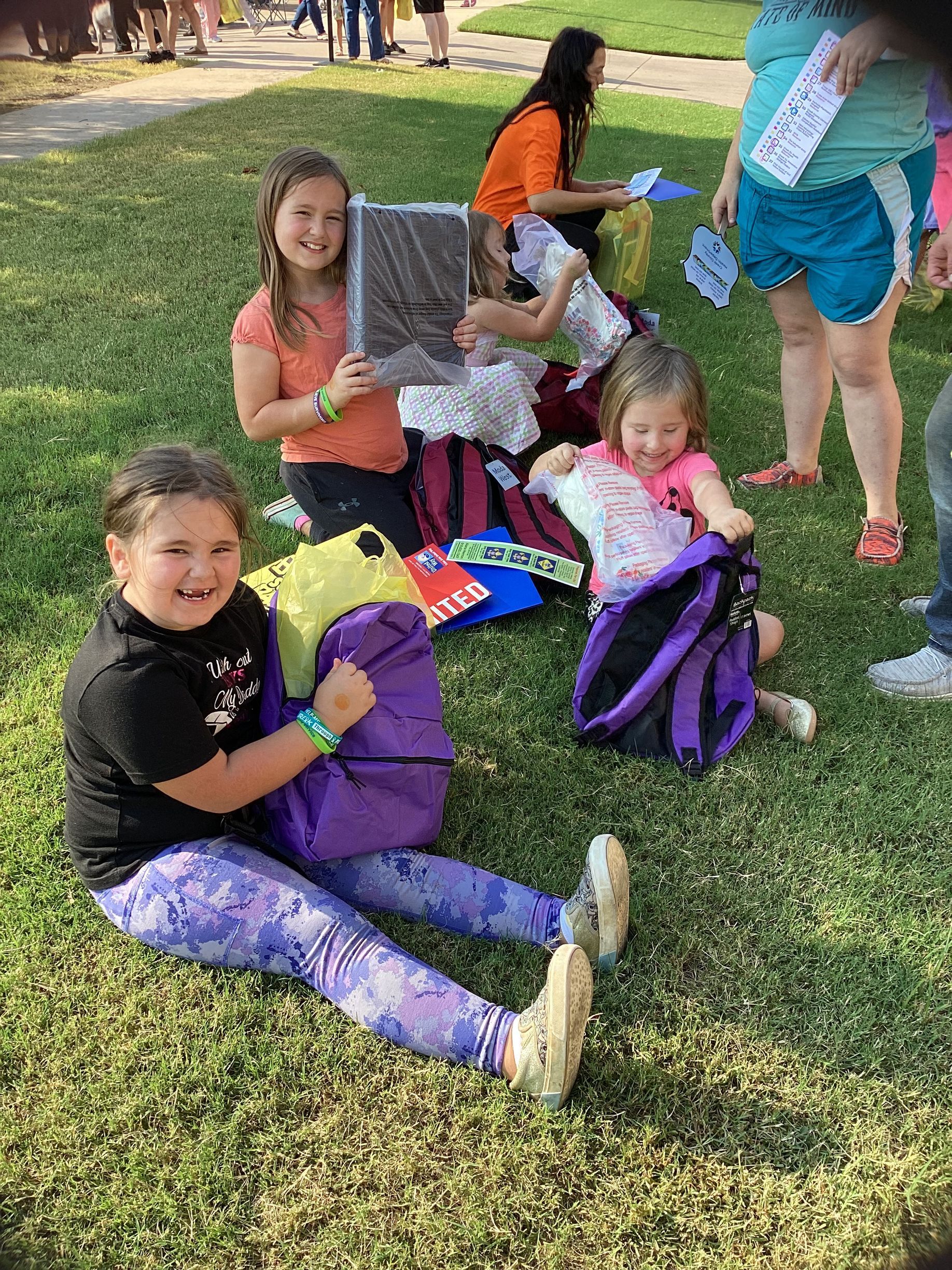 A group of children sitting on grass, some holding bags and supplies, with adults standing nearby on a sunny day.