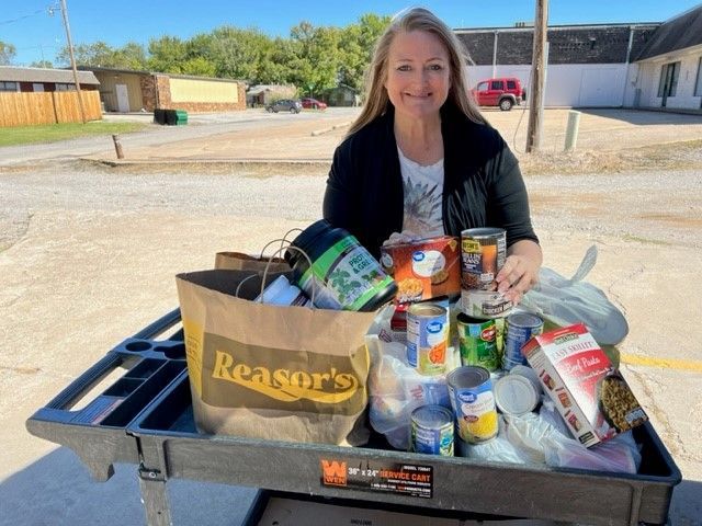 A smiling person holds a can behind a shopping cart filled with grocery bags and canned food in an outdoor parking lot.