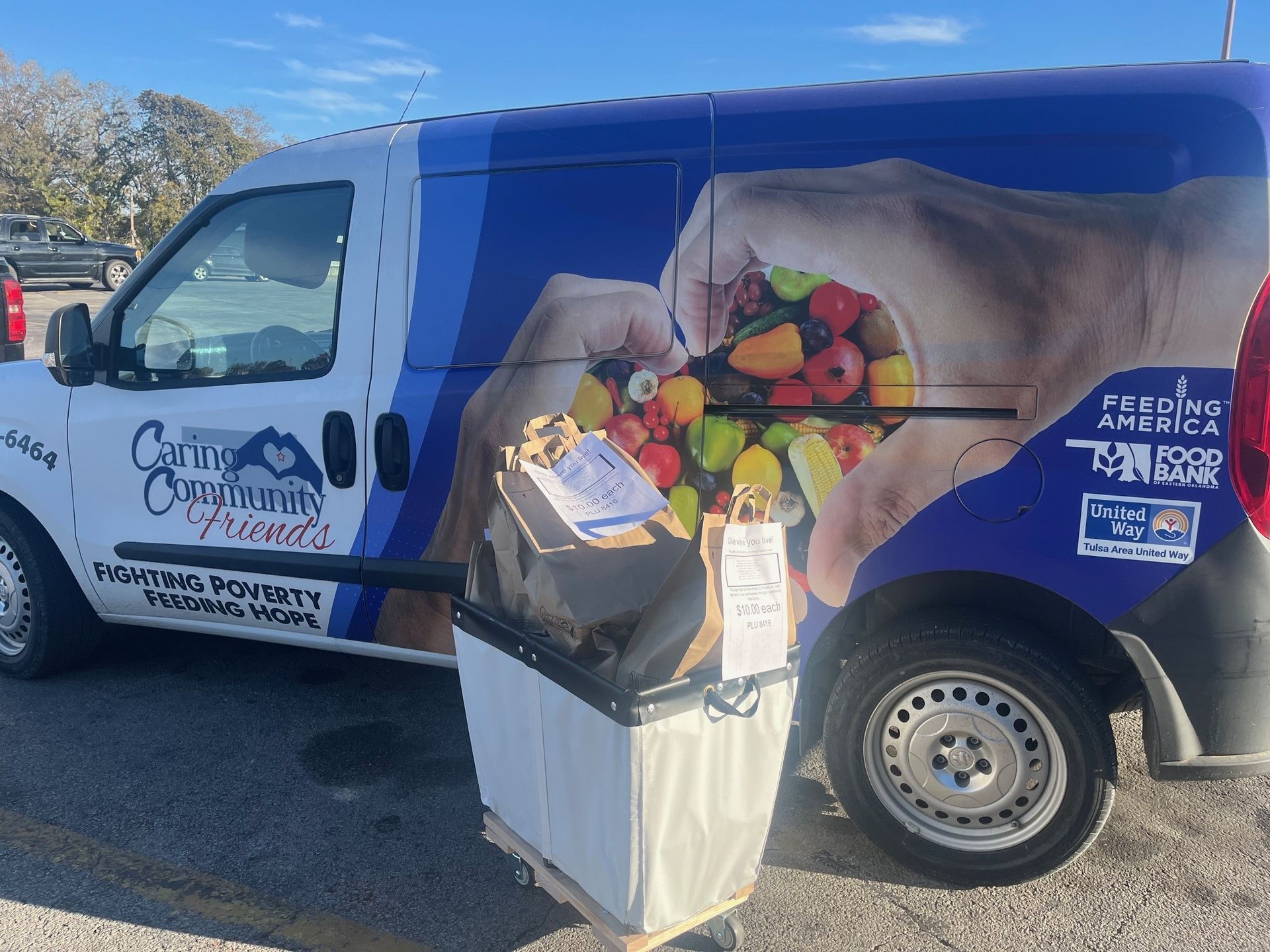 A community food bank van parked outdoors next to a rolling cart filled with bags of food.