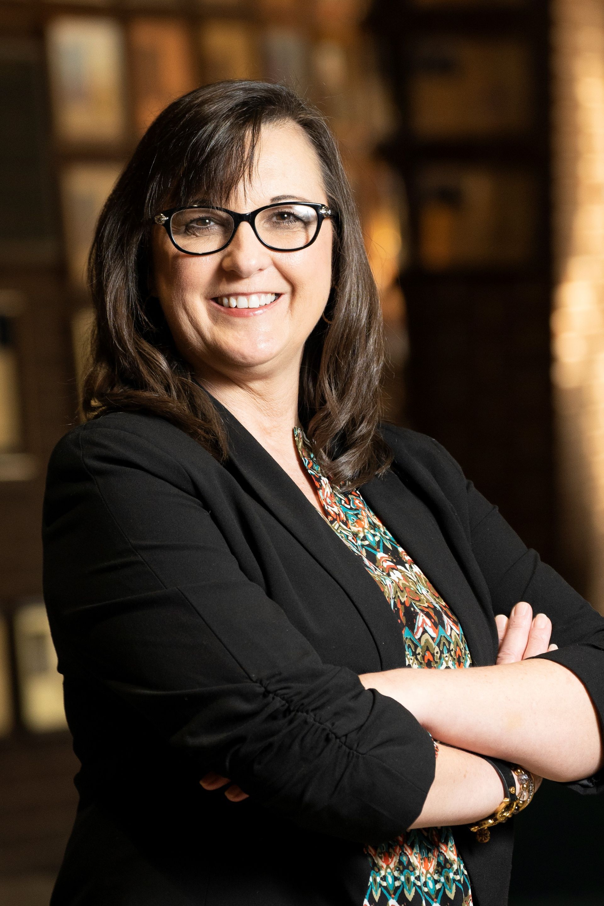 A smiling professional with dark hair and glasses wears a black blazer over a patterned top, arms crossed in a room.