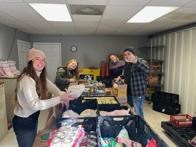 Four people organize food donations into plastic crates and boxes in a well-lit community pantry.