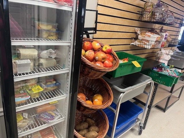 A refrigerated display case filled with packaged food stands next to a three-tiered woven basket stand holding fruit.