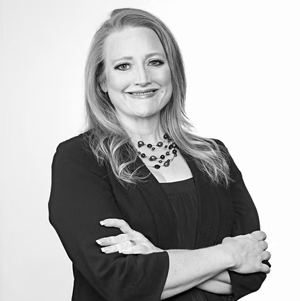 A professional portrait of a smiling person with shoulder-length hair, wearing a dark blazer and necklace, arms crossed.