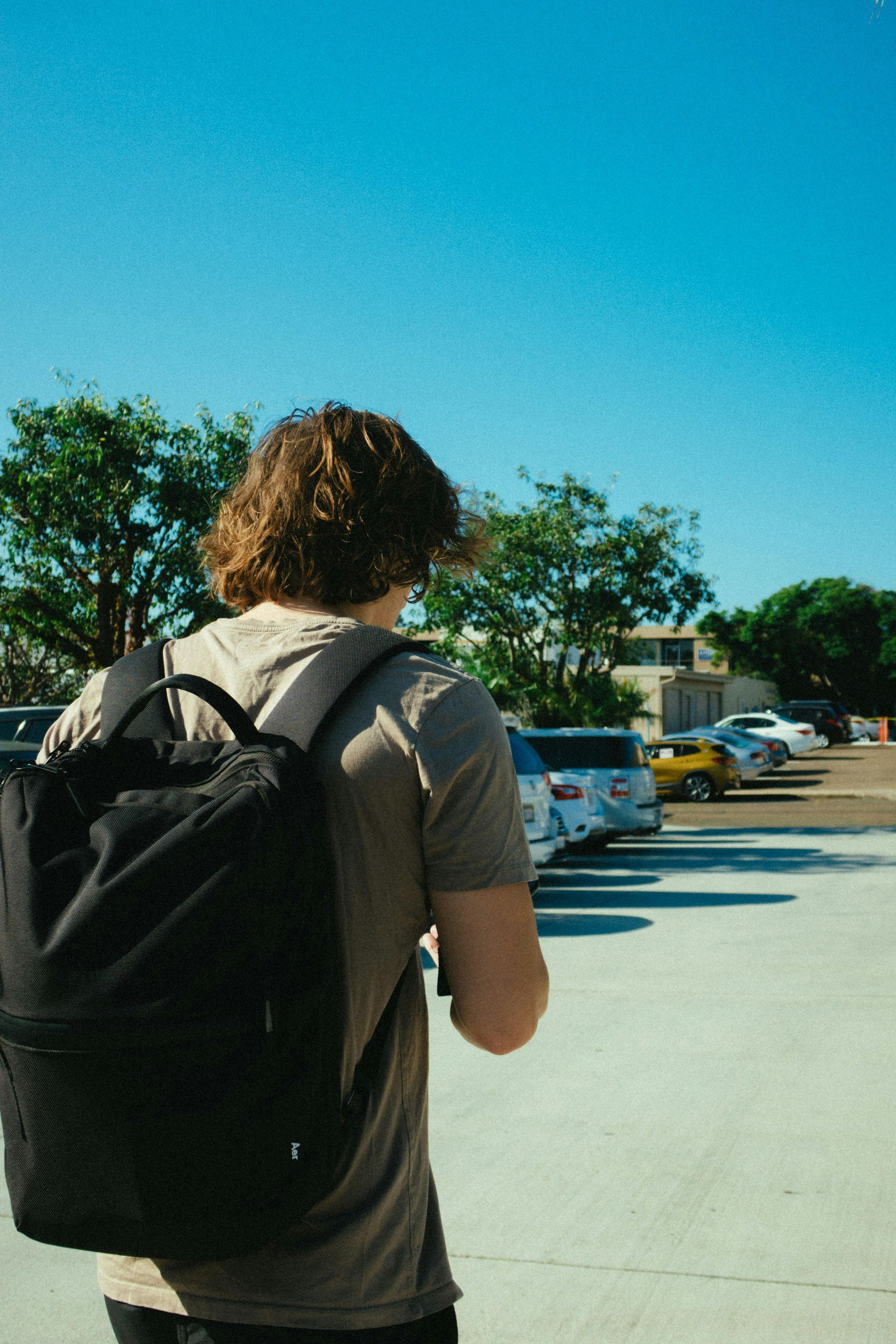 A person with wavy hair wearing a tan t-shirt and a black backpack walks through an outdoor parking lot under a blue sky.