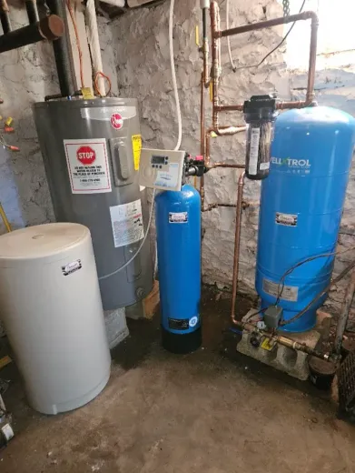 Water heater and filtration system in a basement. Tanks are blue, silver, and white against a stone wall.