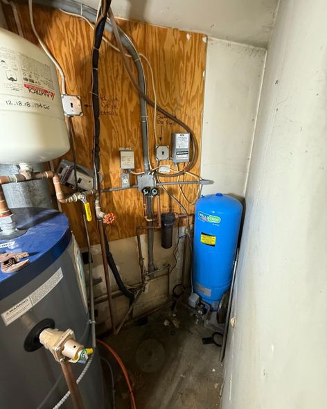 Water heater and pressure tank in a utility room with wooden backing, pipes, and electrical components.