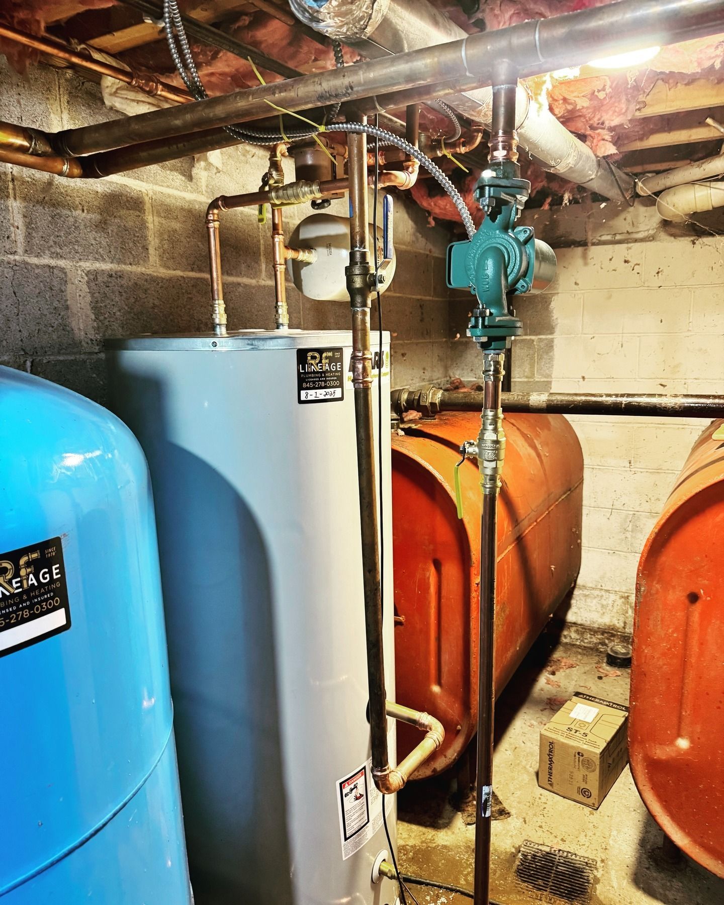 A basement with heating equipment.  A blue water tank is beside a silver tank.  An orange fuel tank stands nearby.
