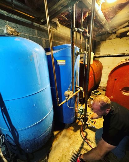 Man working on plumbing in a basement. Blue and red tanks, copper pipes, and exposed brick wall.