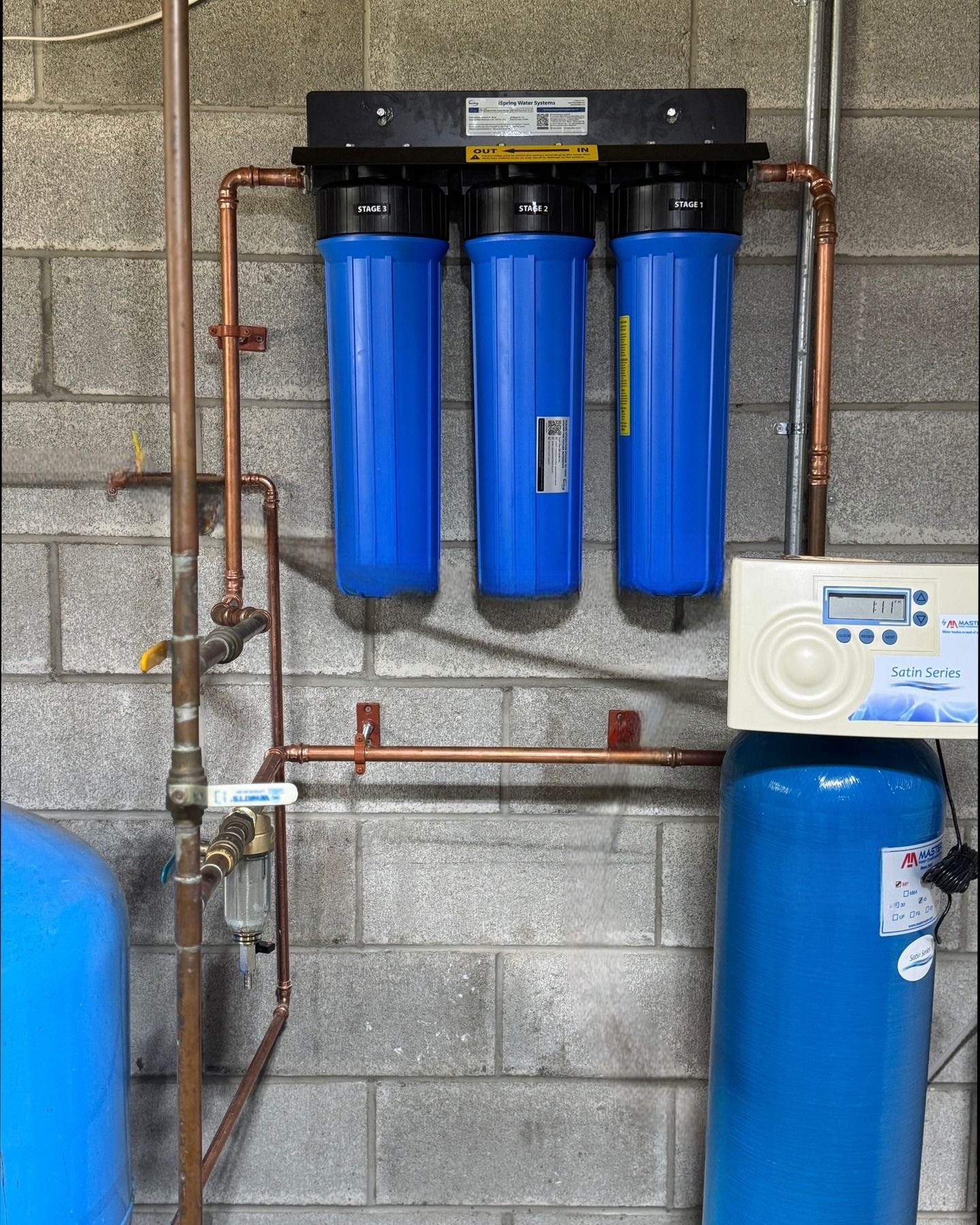 Water filtration system with three blue filters, copper pipes, and control unit on a brick wall.