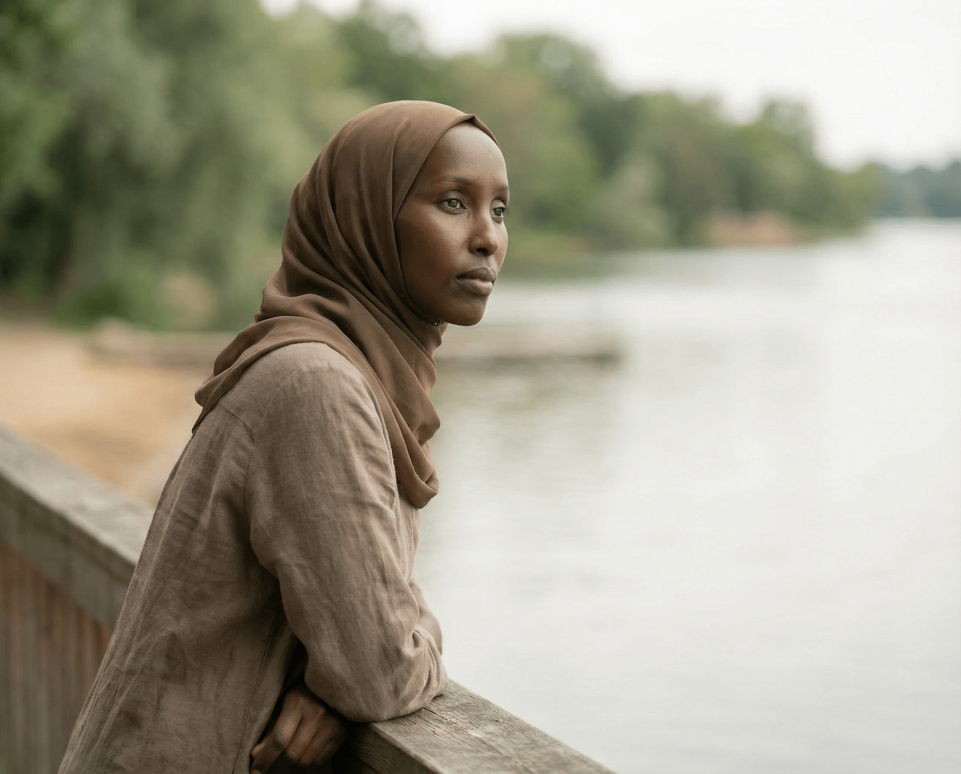 Woman in brown hijab and shirt, leaning on a wooden rail, looking at a river.