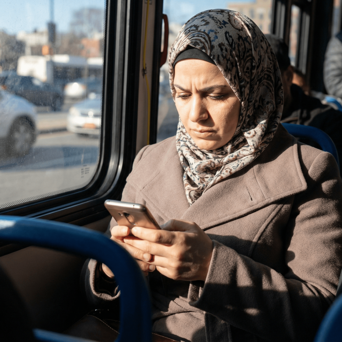 Woman in hijab looking at a phone on a bus. She is wearing a brown coat and sitting by a window.
